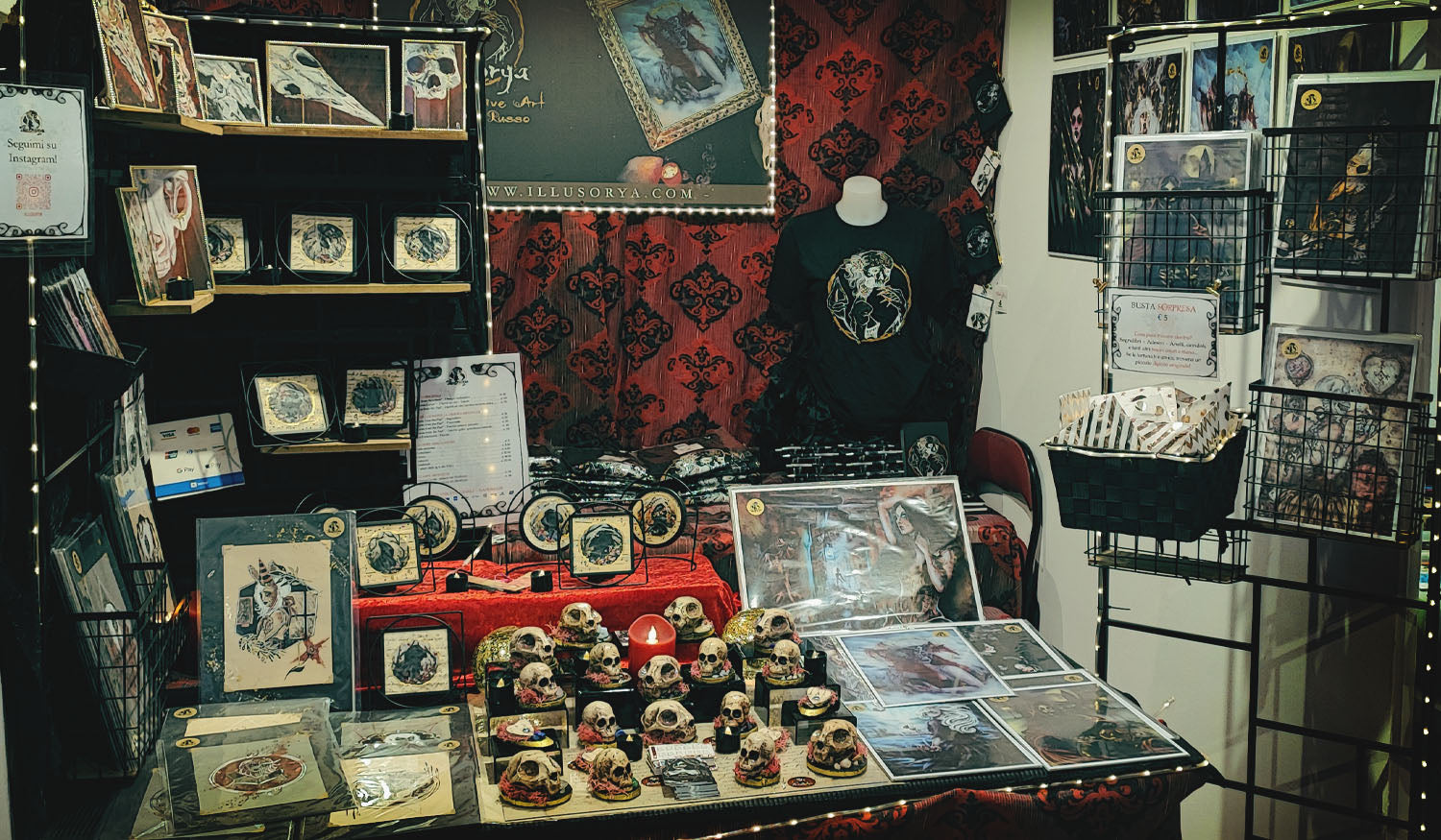 Cover pagina calendario Eventi & Mostre - Foto che rappresenta l'elegante stand goth "Illusorya" in fiera. Rappresentati qui: disegni originali, piccoli dipinti incorniciati, stampe artistiche, decorazioni in ceramica e pietre naturali, t-shirt e gadget con logo stampato. Tema principale: teschi e ritratti oscuri. Fatto a mano, con amore - Stefania Russo, aka Illusorya - Dark Gothic Fantasy Art