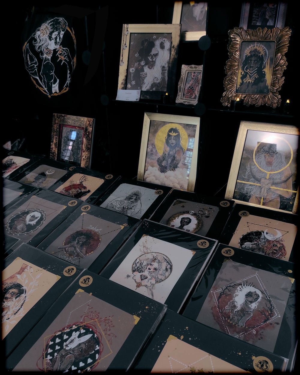 Foto che rappresenta una panoramica della collezione "Tales from Nowhere": disegni goth fatti ad inchiostro nero, bianco, rosso e oro. Sullo sfondo altri piccoli dipinti in acquerello incorniciati e un telo nero con logo stampato - Stefania Russo, aka Illusorya - Dark Gothic Fantasy Art